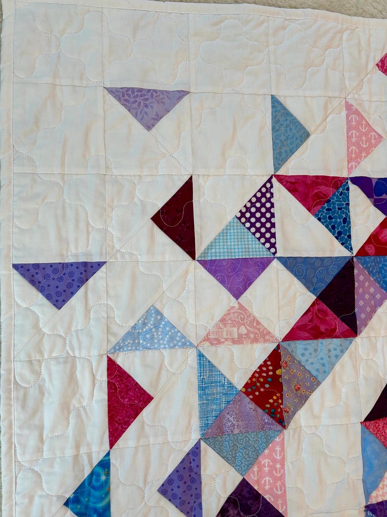 Puede incluir: Primer plano de un edred&oacute;n hecho a mano con un fondo blanco y patrones triangulares coloridos. Los tri&aacute;ngulos son en tonos de azul, morado, rosa y rojo, con varios patrones. El edred&oacute;n est&aacute; cosido a m&aacute;quina con un patr&oacute;n ondulado.