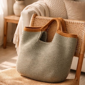 Può includere: Una borsa tote all'uncinetto fatta a mano in una morbida tonalità verde con finiture e manici giallo senape a contrasto. La borsa è su una sedia di vimini, suggerendo un focus sull'arredamento e sugli accessori per la casa.