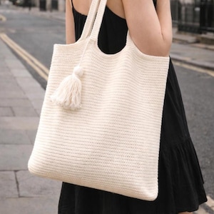 May include: Cream-colored, rectangular tote bag with a tassel. The bag has two thick straps and a large, open top. The bag is made of a woven material, and the tassel is attached to the front. The bag is carried over the shoulder.