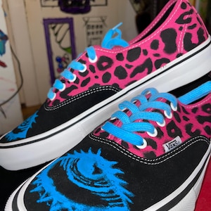 Könnte beinhalten: Paar schwarze und pinkfarbene Sneaker mit Leopardenmuster und blauen Schnürsenkeln. Die Sneaker haben eine weiße Sohle und ein blaues Augendesign auf der Spitze. Das Vans-Logo ist sichtbar.