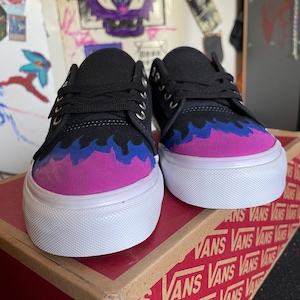 Könnte beinhalten: Ein Paar schwarze Vans-Sneaker mit einem Flammenmuster in Lila, Blau und Rosa. Die Schuhe haben weiße Sohlen und Schnürsenkel. Sie werden auf einem Vans-Schuhkarton präsentiert.