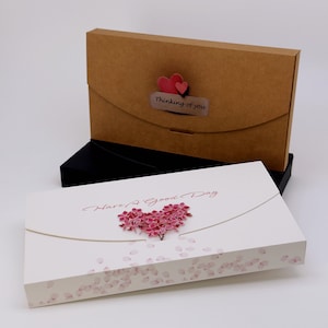 May include: Two gift boxes, one brown and one white, stacked on a black box. The brown box has a flap closure and a sticker that says "Thinking of you" with two red hearts. The white box says "Have a Good Day" with a pink heart.