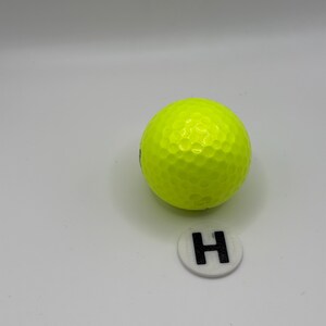Peut inclure: Une balle de golf jaune vif avec une surface à picots. Un petit marqueur circulaire blanc avec un "H" noir est à côté de la balle. Le fond est gris clair uni.