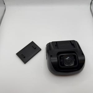 Może przedstawiać: Czarna kamera samochodowa z obiektywem i odłączoną czarną osłoną. Kamera ma prostokątny kształt z zaokrąglonymi krawędziami i małym obiektywem pośrodku. Osłona jest również czarna i prostokątna.