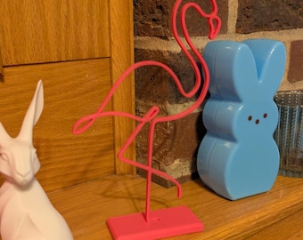 May include: A pink flamingo wire sculpture stands on a pink base, next to a light blue bunny-shaped container. A white rabbit figurine is on the left. The objects are on a wooden surface with a brick background.