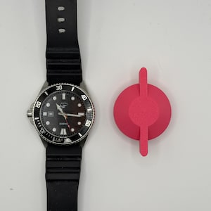 May include: A black wristwatch with a black rubber strap and a silver bezel. The watch face is black with white markings and a date display. A bright pink, circular object with a handle is next to the watch.