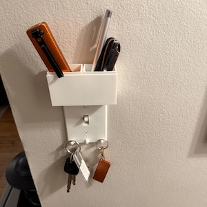 Puede incluir: Un organizador de pared blanco impreso en 3D con un diseño de interruptor de luz. Cuenta con dos compartimentos que contienen bolígrafos y un llavero con dos llaveros. El organizador está montado en una pared de color claro.