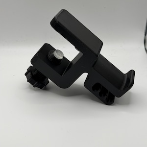 May include: A black, 3D-printed clamp with a rectangular top and a knob. The clamp has a hinged design and a clear, circular fastener. The object is set against a white background.