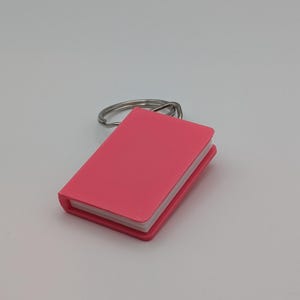 May include: A small, pink, miniature book keychain with a silver-colored key ring. The book has a smooth, solid pink cover and white pages. The keychain is a simple, yet charming accessory.