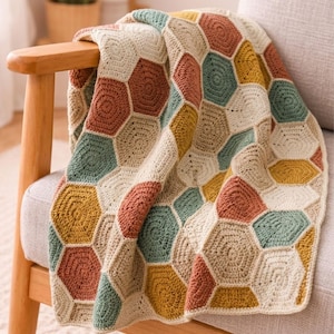 Puede incluir: Una manta de ganchillo hecha a mano, colocada sobre una silla. La manta presenta un patrón de panal en tonos crema, marrón, amarillo y verde. La manta está hecha de hilo suave y parece acogedora e invitadora.