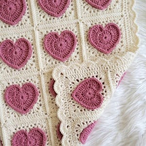 May include: A handmade crochet blanket with a pattern of pink hearts on a cream background. The blanket has a scalloped edge and is made of soft yarn. The hearts are evenly spaced across the blanket. The blanket is made with a soft yarn.