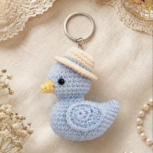 May include: A light blue crocheted duck keyring featuring a small cream-coloured straw-style hat, a yellow beak, and black bead eyes. The duck is attached to a circular silver-toned metal split ring for keys, showcasing detailed yarn stitching.
