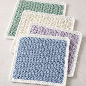May include: A set of four square crochet washcloths featuring a textured stitch pattern. Each washcloth has a solid colored center in sage green, cream, lavender, or dusty blue, and is finished with a solid white crocheted border edge.