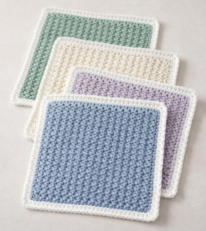 May include: A set of four square crochet washcloths featuring a textured stitch pattern. Each washcloth has a solid colored center in sage green, cream, lavender, or dusty blue, and is finished with a solid white crocheted border edge.