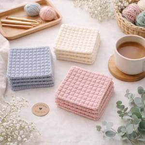 May include: Stacks of handmade crochet coasters in pastel colors: light blue, cream, and pink. The coasters have a textured, square design. A wooden tray with crochet hooks and yarn balls, a cup of coffee, and a small sign that says "handmade with love" are also in the image.