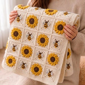 Peut inclure: Une couverture au crochet de couleur crème avec un motif répété de tournesols jaunes et de bourdons. La couverture est composée de panneaux carrés, chacun avec un tournesol ou une abeille. Les tournesols ont des centres bruns. La couverture est pliée.