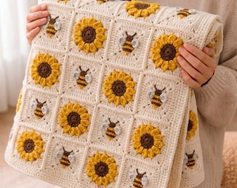 May include: A handmade crochet blanket featuring a repeating square pattern of yellow and brown sunflowers alternating with small bees with white wings, all set against a cream-colored yarn background with a textured, knotted stitch design.