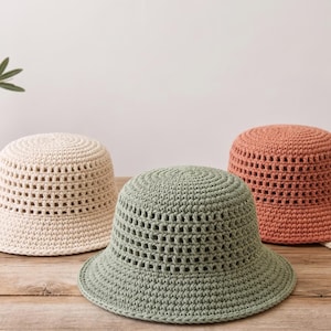 May include: Three crochet bucket hats in cream, sage green, and terracotta orange. Each hat features a solid crown and a wide, open-weave mesh pattern around the sides, finished with a flat, circular top and a short, downward-sloping brim.