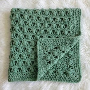 Peut inclure: Une couverture en crochet vert sauge présentant un motif texturé en points noisette disposé selon un design géométrique répétitif. La couverture possède une bordure unie et est repliée sur un coin pour révéler le travail en relief.