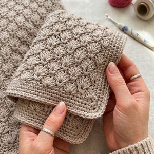 Puede incluir: Una tela tejida a ganchillo de color beige con un patrón repetitivo de puntos de concha y una superficie texturizada. El tejido está doblado, mostrando una disposición densa y uniforme de los puntos. Un ganchillo y un carrete de hilo son visibles al fondo.