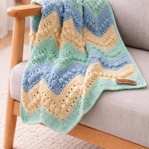 Può includere: Una coperta all'uncinetto fatta a mano con un motivo a zig-zag, drappeggiata su una sedia. La coperta presenta strisce alternate di azzurro, crema e verde menta. La trama include punti in rilievo. Una piccola etichetta recita "Hugs & Kisses".
