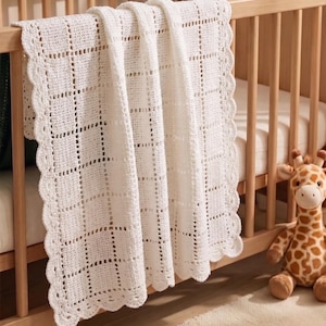 Puede incluir: Una manta de bebé blanca, tejida a ganchillo, con un patrón de rejilla y bordes festoneados, sobre una cuna de madera. Un juguete de peluche de jirafa está junto a la cuna. La manta está hecha de hilo suave y texturizado.