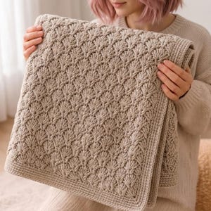 Puede incluir: Una manta beige tejida a mano con un patrón de hojas repetido. La manta es rectangular y tiene un borde grueso y texturizado. El color suave y neutro y el diseño intrincado la convierten en un elemento de decoración del hogar acogedor.