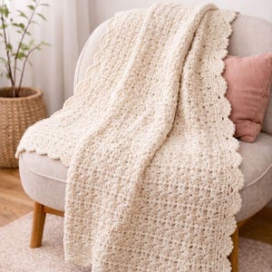 May include: A cream-colored crochet blanket featuring a repeating star-like textured pattern throughout the center and a decorative scalloped edge border. The textile is draped over a chair, showcasing the intricate yarn work and soft neutral tone.