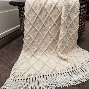 Puede incluir: Manta de color crema, tejida a mano con ganchillo, con un patrón de diamantes y flecos. La manta está sobre una cesta de mimbre marrón oscuro. El patrón de diamantes se crea con puntos en relieve, agregando textura.