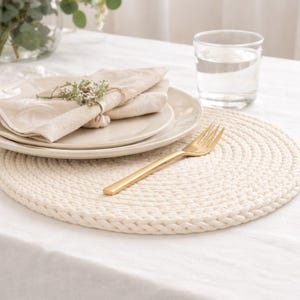 Peut inclure: Un set de table rond tressé de couleur crème avec une fourchette dorée, des assiettes et une serviette pliée. La serviette est attachée avec de la ficelle et décorée de verdure. Un verre d'eau et un vase de fleurs sont également sur la nappe blanche.