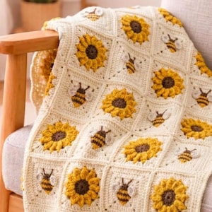 May include: A handmade crochet blanket featuring a repeating pattern of square motifs. The design alternates between textured yellow and brown sunflower squares and cream-colored squares decorated with small, embroidered bumblebee appliques.