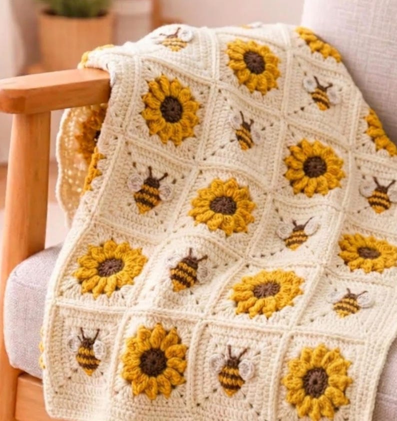 May include: A handmade crochet blanket featuring a repeating pattern of square motifs. The design alternates between textured yellow and brown sunflower squares and cream-colored squares decorated with small, embroidered bumblebee appliques.