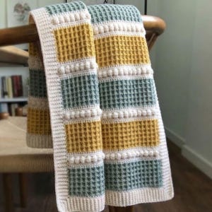 Puede incluir: Una manta de ganchillo con un patrón de punto de gofre en bloques alternos de color amarillo mostaza y verde salvia. La manta está bordeada por un borde de color crema e incluye filas horizontales de puntos de burbuja en relieve.