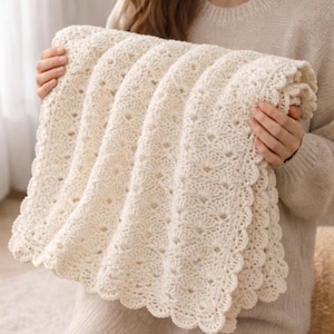 May include: Cream-colored, crocheted blanket with a scalloped edge. The blanket features a detailed openwork pattern. The soft, textured blanket is folded and held, showcasing its intricate design. A cozy, handmade item.