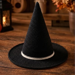 Puede incluir: Un sombrero de bruja de ganchillo negro con una corona alta y puntiaguda y un ala circular ancha. La base de la corona presenta una banda decorativa de ganchillo blanco que rodea el sombrero, creando un contraste con el hilo negro.