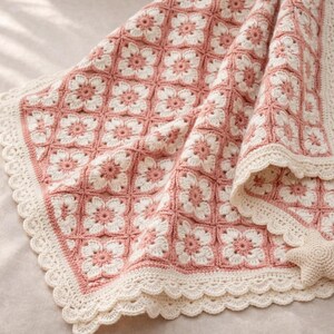 May include: A handmade crochet blanket featuring a floral pattern in shades of pink and cream. The blanket has a scalloped edge and is made with a soft yarn. The design is a repeating pattern of flowers.