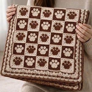 Può includere: Una coperta quadrata all'uncinetto con un motivo a scacchiera di quadrati alternati marrone scuro e color crema. Ogni quadrato presenta un motivo centrale a forma di impronta di zampa. La coperta è rifinita con un bordo decorativo all'uncinetto color crema.