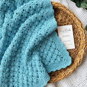 Può includere: Una coperta per neonati all'uncinetto blu chiaro con un motivo a punto in rilievo strutturato. La coperta è piegata su un vassoio intrecciato, con un'etichetta che recita "Tiny Baby Collection" e "100% Cotone organico."