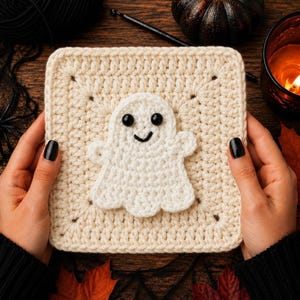 May include: A square crochet granny square in cream yarn featuring a centered white ghost applique. The ghost has two black plastic safety eyes and a stitched black mouth. The square has a textured stitch pattern with small dark accents at the corners.