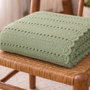 Könnte beinhalten: Eine gefaltete, salbeigrüne Strickdecke mit einem gewellten Rand liegt auf einem Korbstuhl. Die Decke hat Reihen von kleinen, gleichmäßig verteilten Löchern, die dem weichen, strukturierten Stoff ein dekoratives Element verleihen. Die Decke ist ordentlich gefaltet.