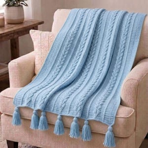 Puede incluir: Una manta de punto de cable azul claro con borlas, sobre un sillón beige. La manta presenta un patrón de cable repetido y borlas azules a juego en el borde inferior. Un artículo de decoración del hogar suave y acogedor.