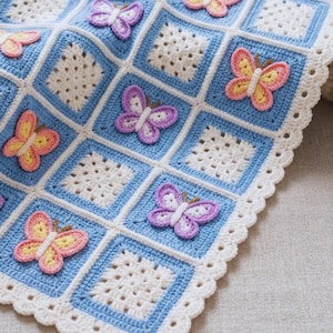 May include: A handmade crochet blanket featuring a grid of alternating white square motifs and light blue square motifs. The blanket is decorated with three-dimensional crocheted butterflies in shades of pink, yellow, and purple, finished with a scalloped white border.