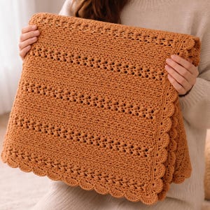 May include: A folded, crocheted blanket in a warm, earthy orange hue. The blanket features a detailed openwork pattern with scalloped edges. The texture is visible, suggesting a soft, handmade item, perfect for home decor.