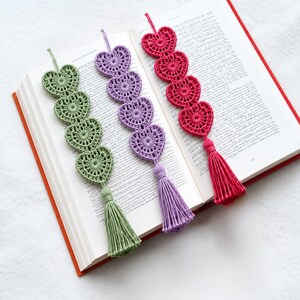 Peut inclure: Trois marque-pages en crochet faits main, de couleur vert sauge, lavande et rose vif. Chaque marque-page présente une chaîne verticale de quatre motifs en forme de cœur interconnectés avec des dentelles, terminés par un pompon assorti.