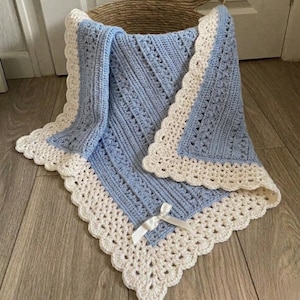 Könnte beinhalten: Eine hellblaue Häkeldecke mit einem strukturierten Rippenmuster und einem breiten, cremefarbenen, wellenförmigen Rand. Die Decke ist mit einer kleinen, weißen Satinschleife am unteren Rand verziert, die eine filigrane Handarbeit aufweist.