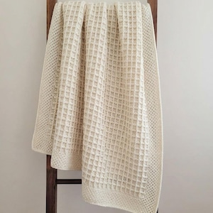 May include: Cream-coloured, waffle-knit blanket draped over a wooden ladder. The blanket has a textured, square pattern and a solid border. The neutral colour and simple design suggest a cosy, home decor item.