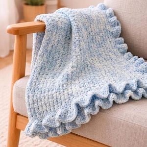 May include: A soft, hand-crocheted blanket featuring a variegated pattern of light blue and white yarn. The blanket has a textured, woven stitch throughout and is finished with a decorative, ruffled edge that runs along the side of the fabric.