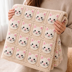 May include: A cream-colored crochet blanket featuring a repeating grid pattern of individual square blocks. Each block contains a circular, white cat face motif with pink ears, black embroidered eyes, a pink nose, and black whiskers.