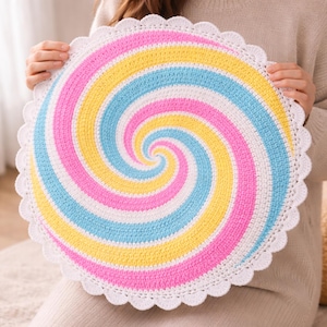 Könnte beinhalten: Eine runde, gehäkelte Matte mit einem Spiralmuster in den abwechselnden Farben Weiß, Pastellrosa, Gelb und Hellblau. Das kreisförmige Stück ist mit einem dekorativen weißen Wellenrand versehen, was für eine strukturierte Optik sorgt.