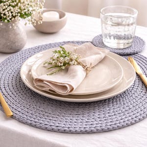 May include: A table setting featuring a light blue crocheted placemat, two white plates, a folded beige napkin, and gold cutlery. A small vase with white flowers and a glass of water complete the elegant dining arrangement.
